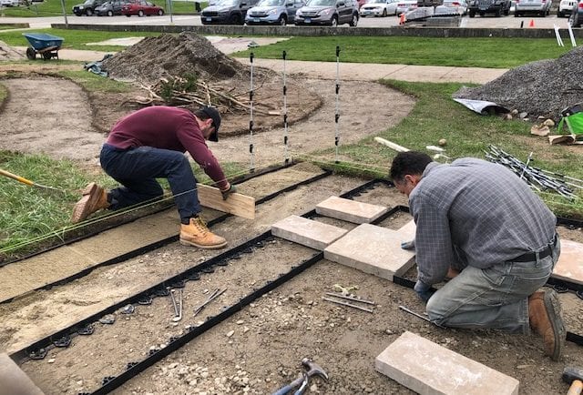 Jacob uses a screed board to level the sand in the edging channel while Charlie checks the fit of a paver